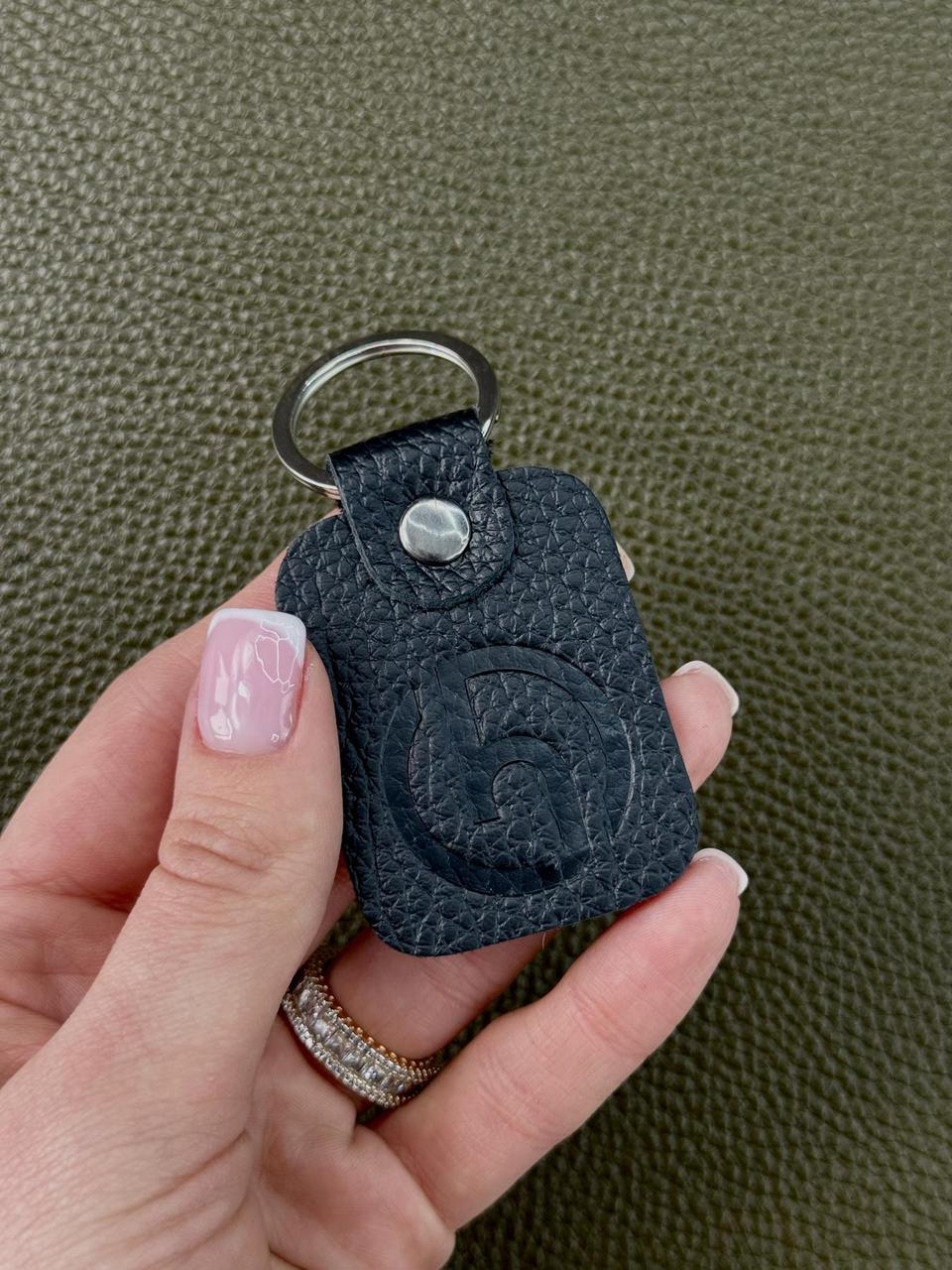 A hand holding a small black textured leather keychain with a silver metal key ring and snap button, set against a dark olive green textured background.