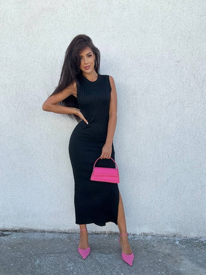 Woman in a black sleeveless dress holding a pink handbag against a white wall.