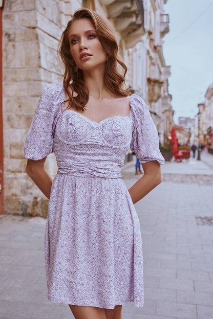 Woman in a light purple floral dress standing on a street with buildings in the background