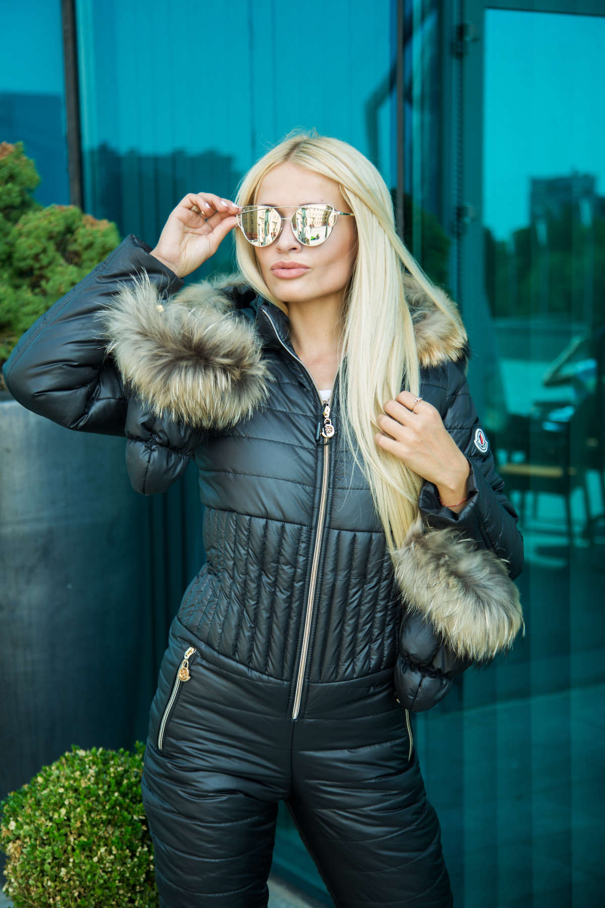 Woman in black quilted jacket with fur trim adjusting sunglasses against a reflective glass background