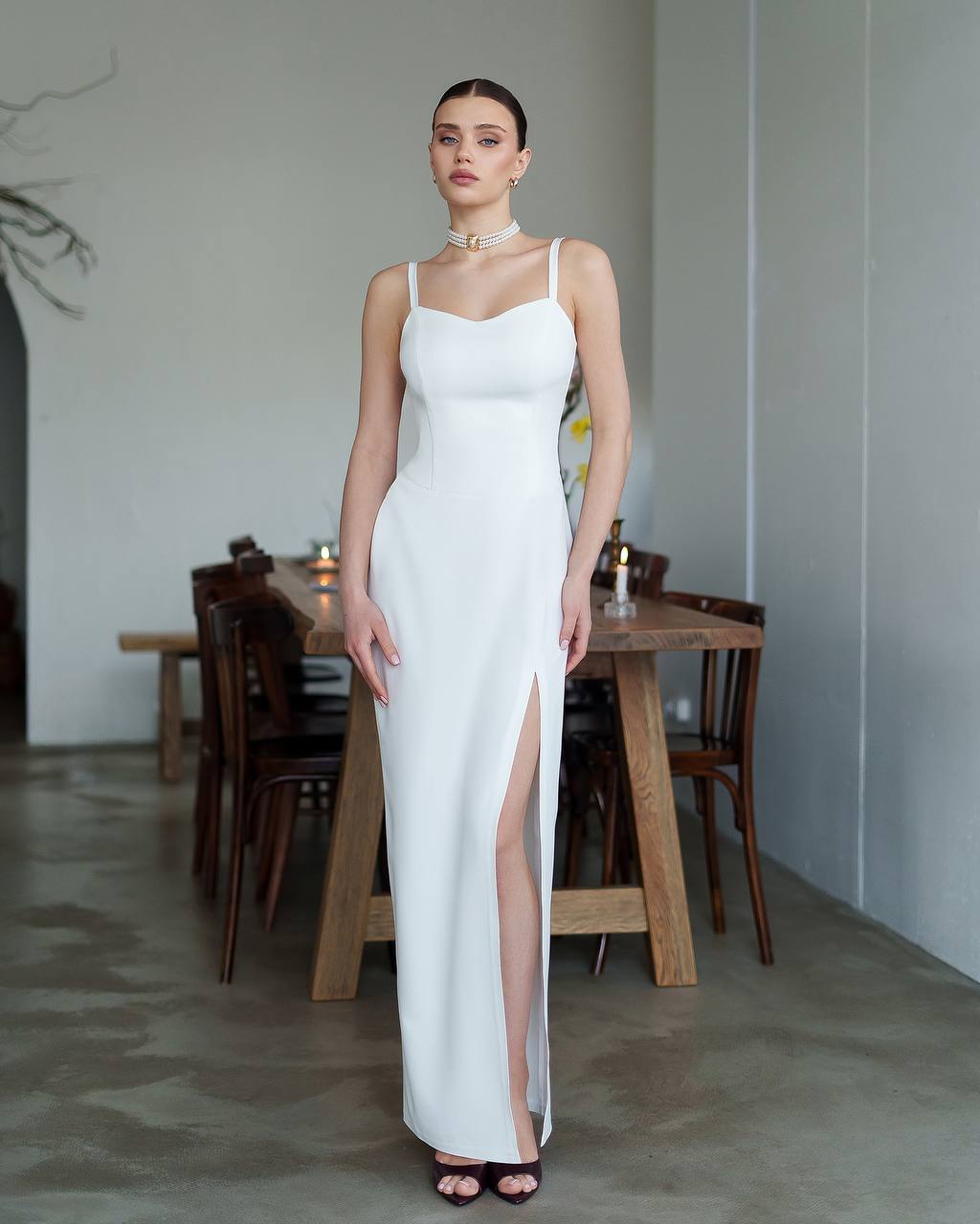 Woman in a white dress standing in a room with wooden furniture.
