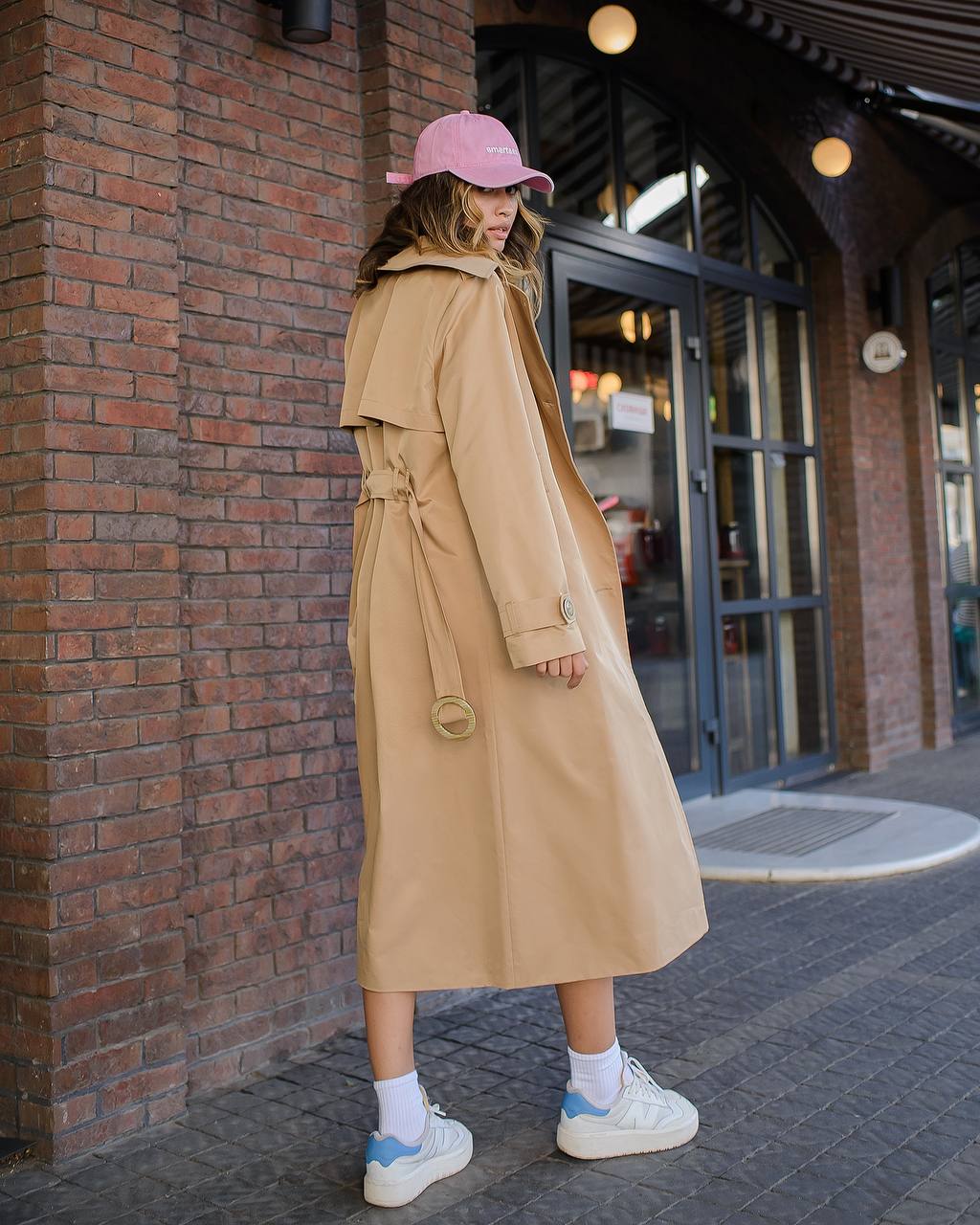 Person wearing a beige trench coat and pink cap standing against a brick wall.