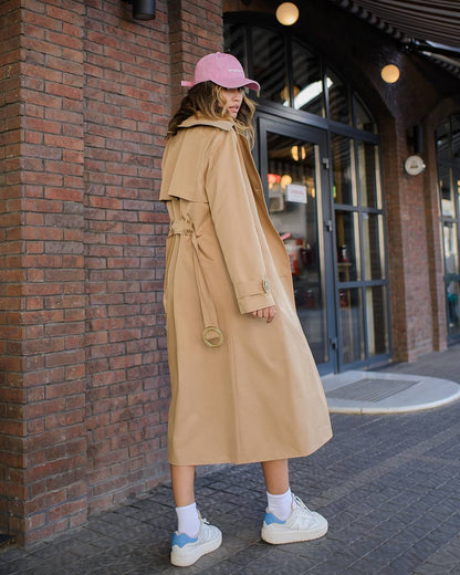 Person wearing a beige trench coat and pink cap standing against a brick wall.