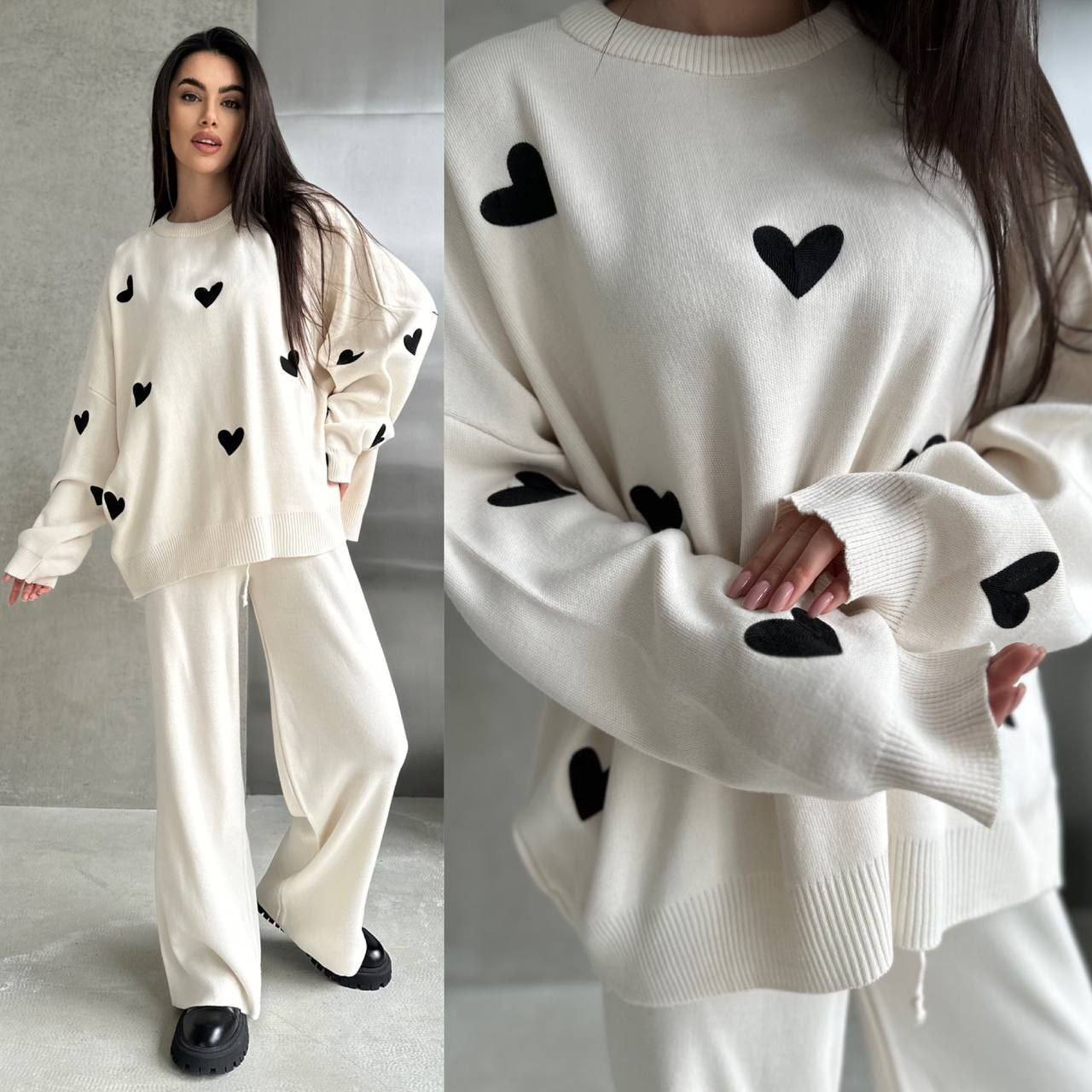 White sweater with black heart patterns worn by a person, with close-up details on a neutral background.