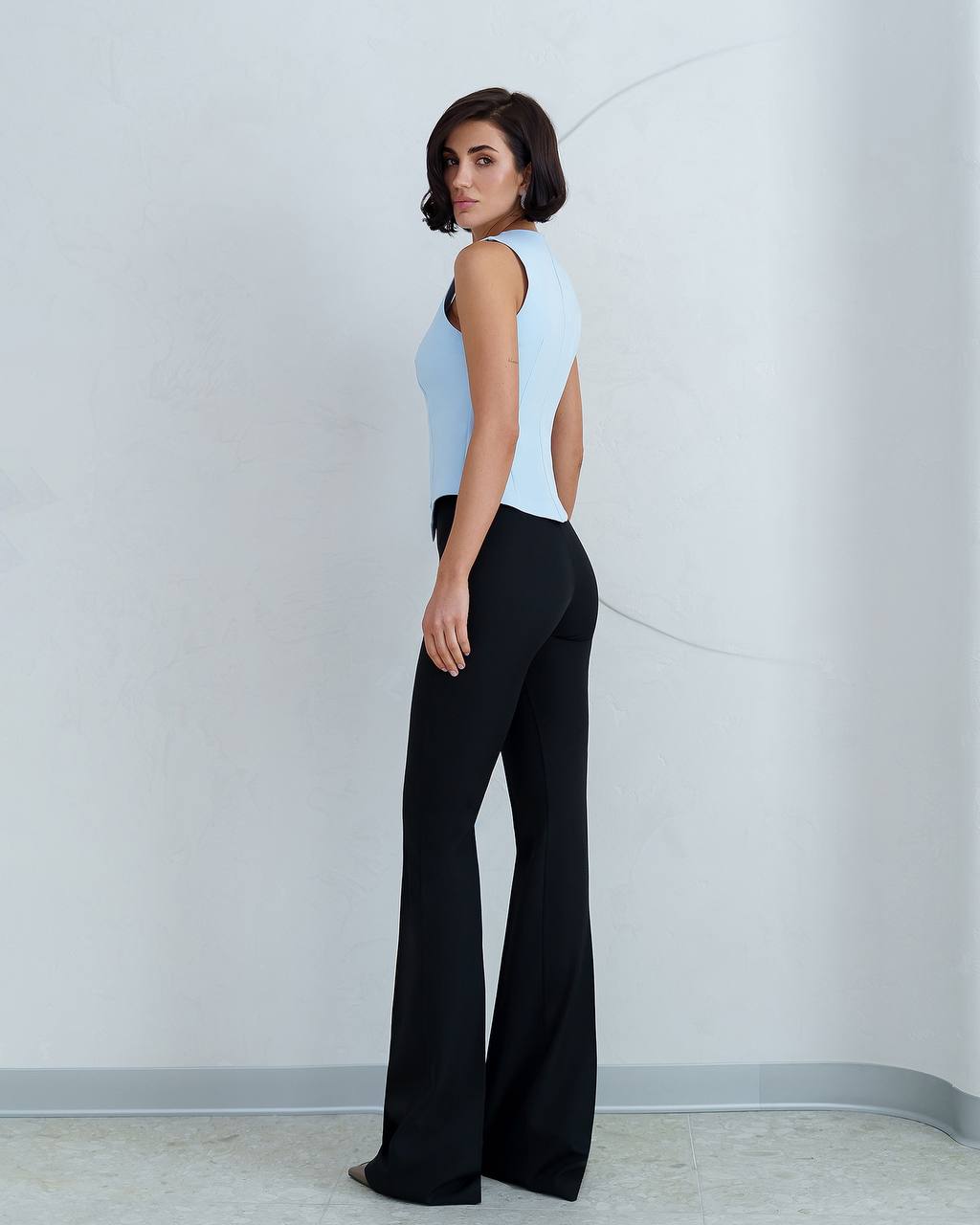 Woman wearing a light blue sleeveless top and black flared pants against a white wall.