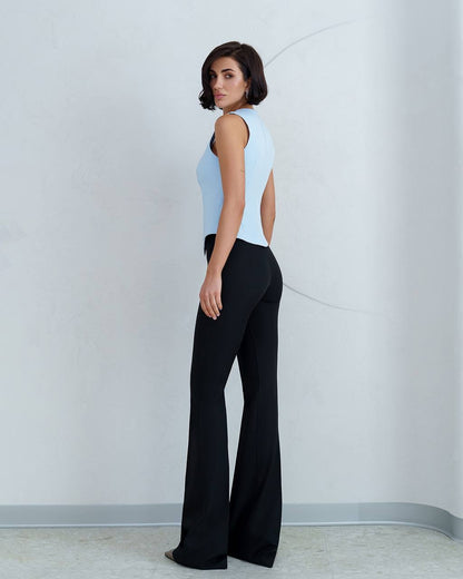 Woman wearing a light blue sleeveless top and black flared pants against a white wall.