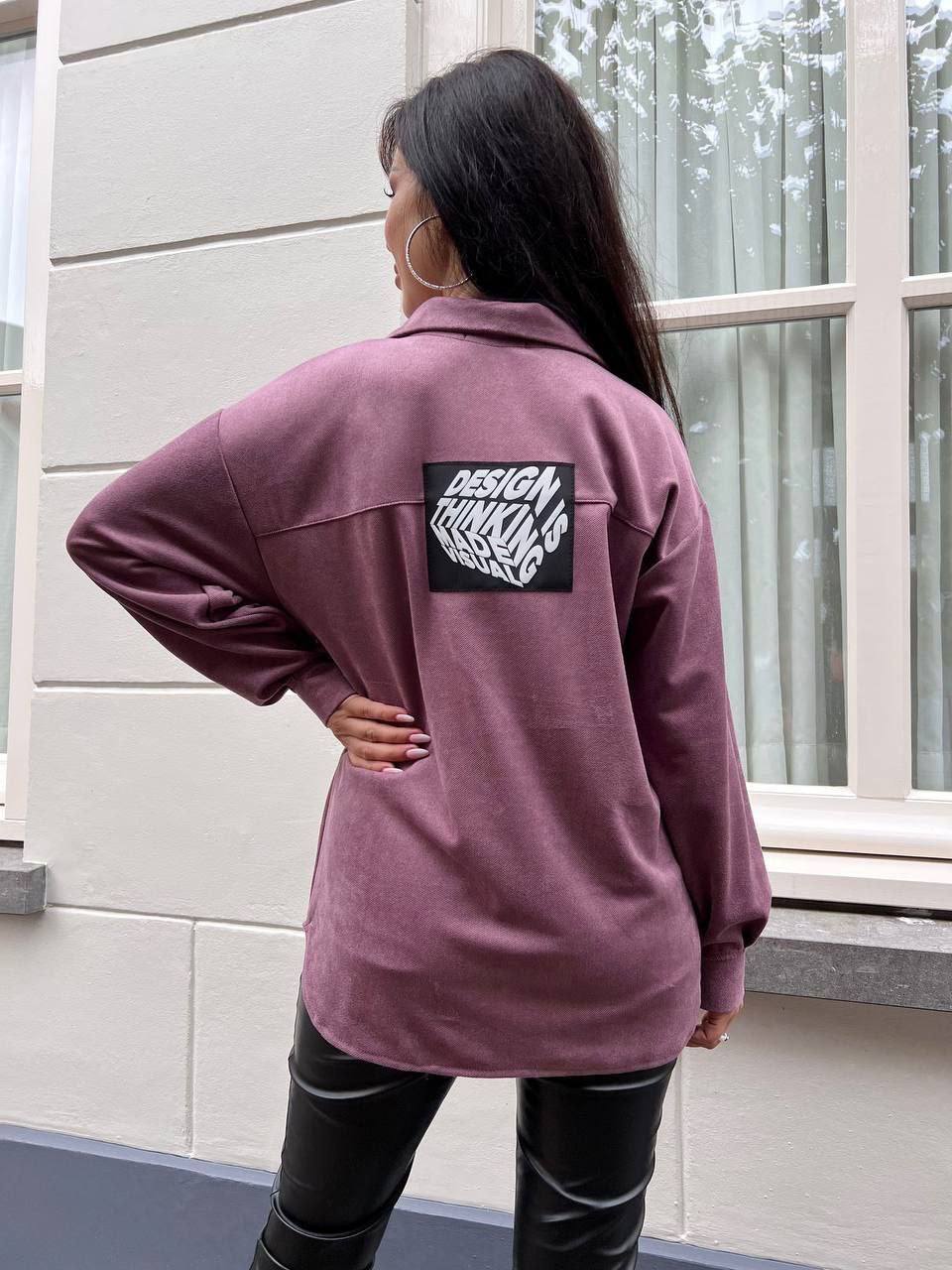 Person wearing a purple jacket with a visible brand logo, standing in front of a window.