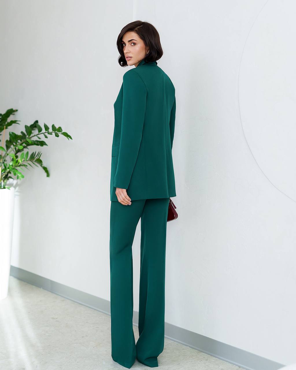 Woman in a green suit standing against a white wall with a plant to the left.