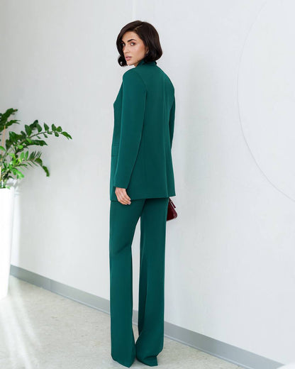 Woman in a green suit standing against a white wall with a plant to the left.