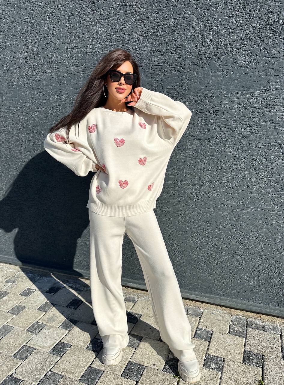 Woman wearing a white sweatshirt with heart patterns and white pants against a gray wall.