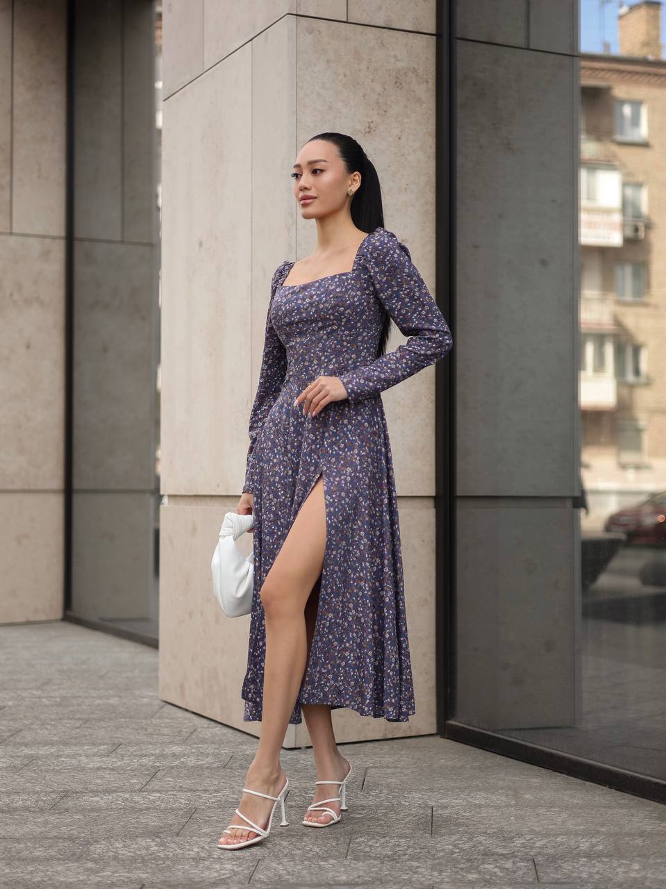 Woman in a purple floral dress standing against a building corner.