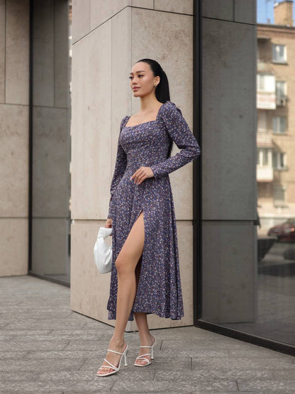 Woman in a purple floral dress standing against a building corner.