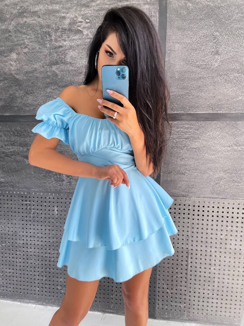 Woman wearing a light blue off-shoulder dress taking a mirror selfie against a gray wall.