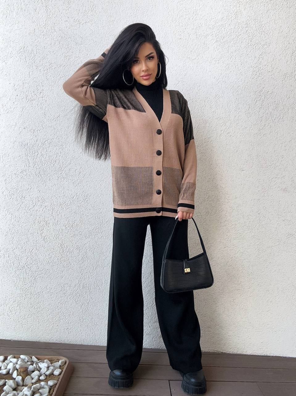 Woman wearing a beige and black cardigan with black pants, holding a black handbag against a white wall.