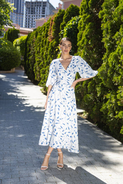 Woman in a white dress with blue patterns standing on a sidewalk with greenery in the background
