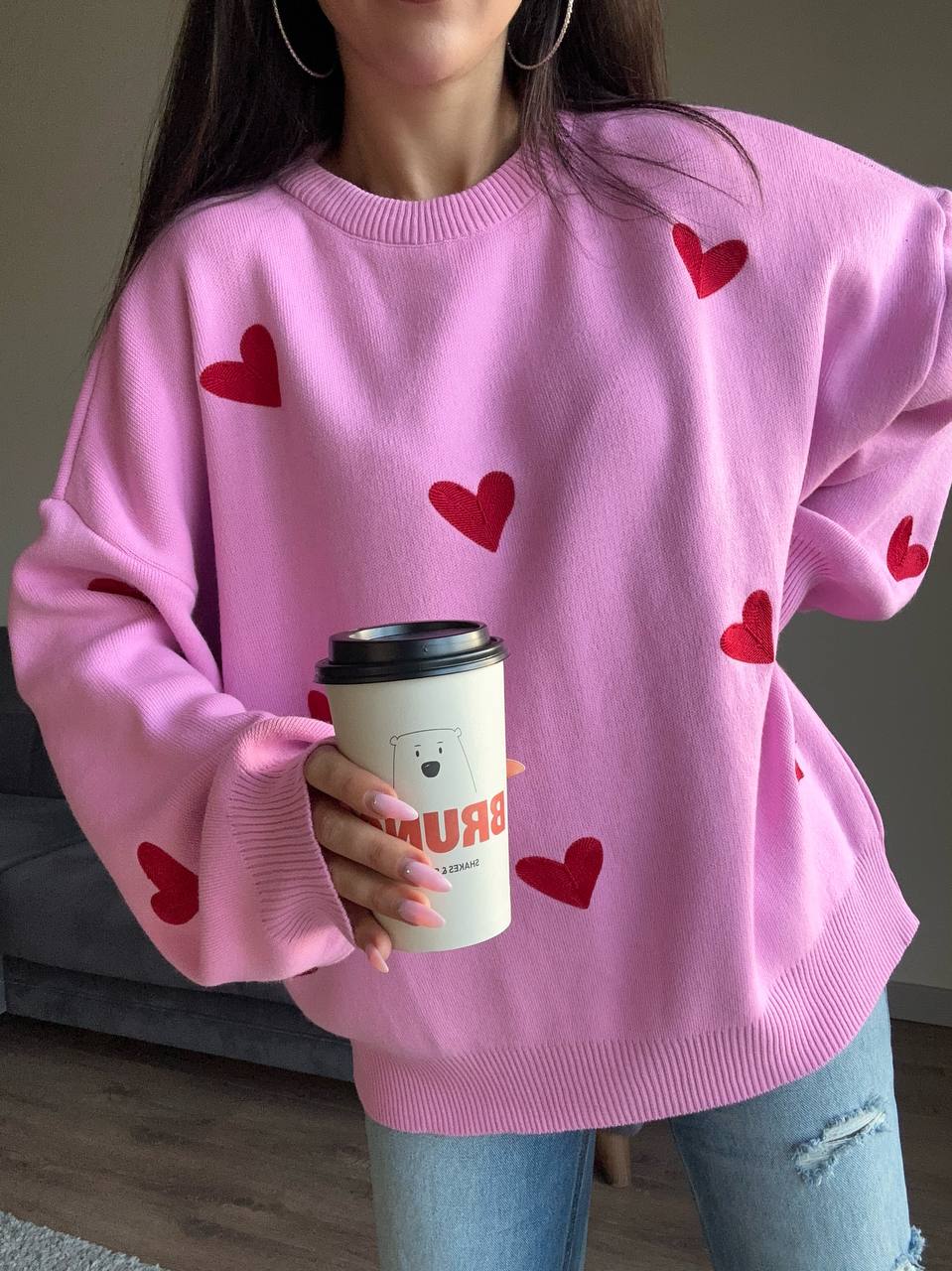 Person wearing a pink sweater with red hearts and holding a coffee cup.