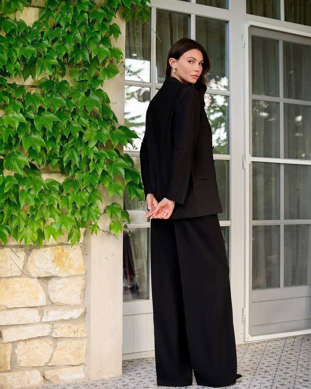 Woman in a black outfit standing in front of a stone wall with greenery