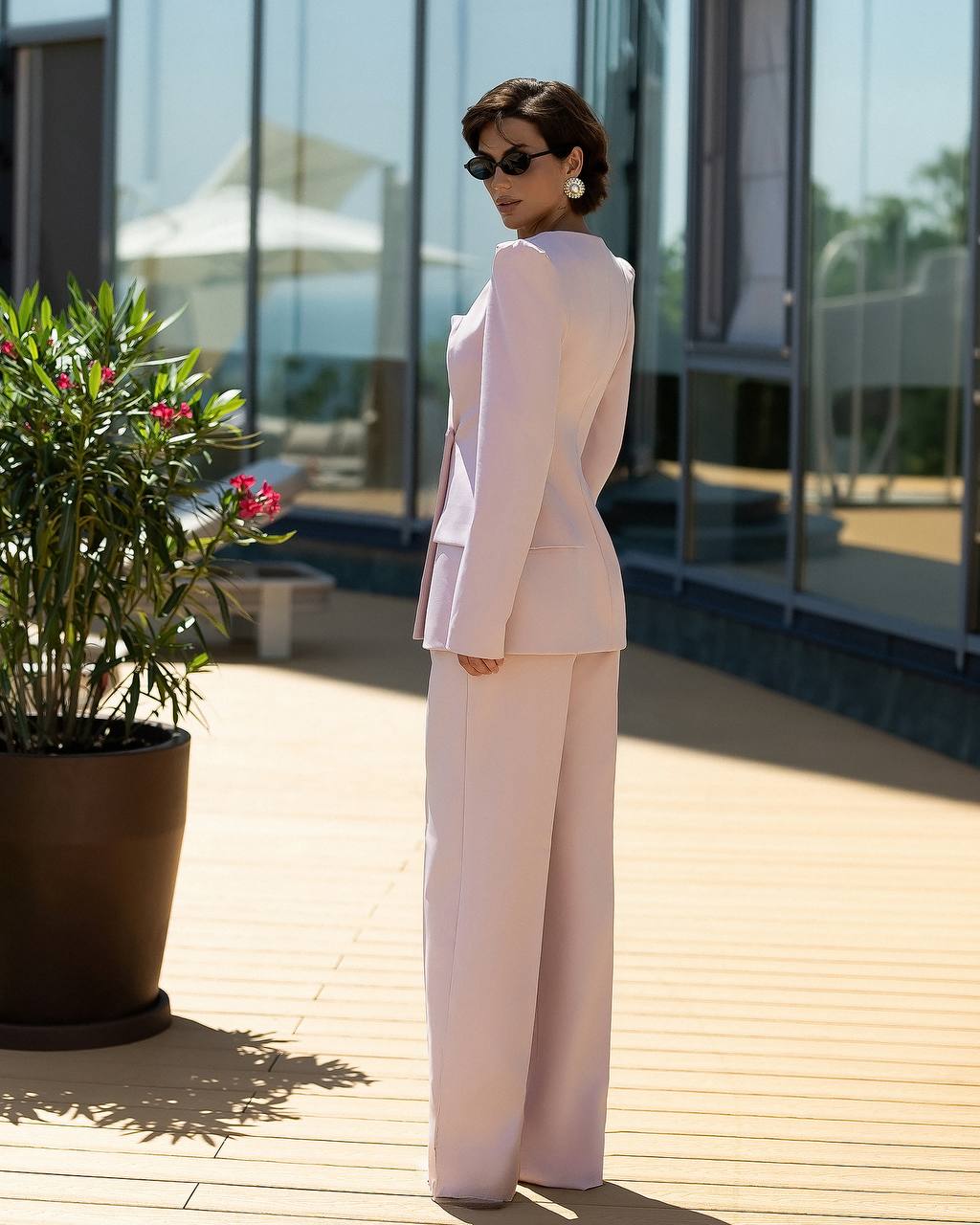 A woman stands on a sunlit outdoor deck wearing a light blush pink tailored suit with a structured blazer and wide-leg trousers. She is photographed from the back, with large windows, a potted plant, and reflections visible in the background.