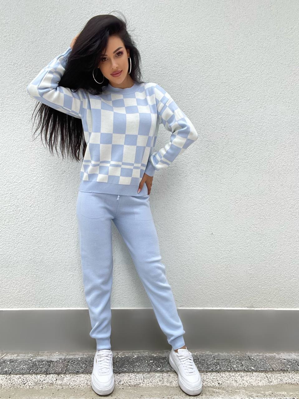 Woman wearing a checkered sweater and light blue pants against a gray wall.