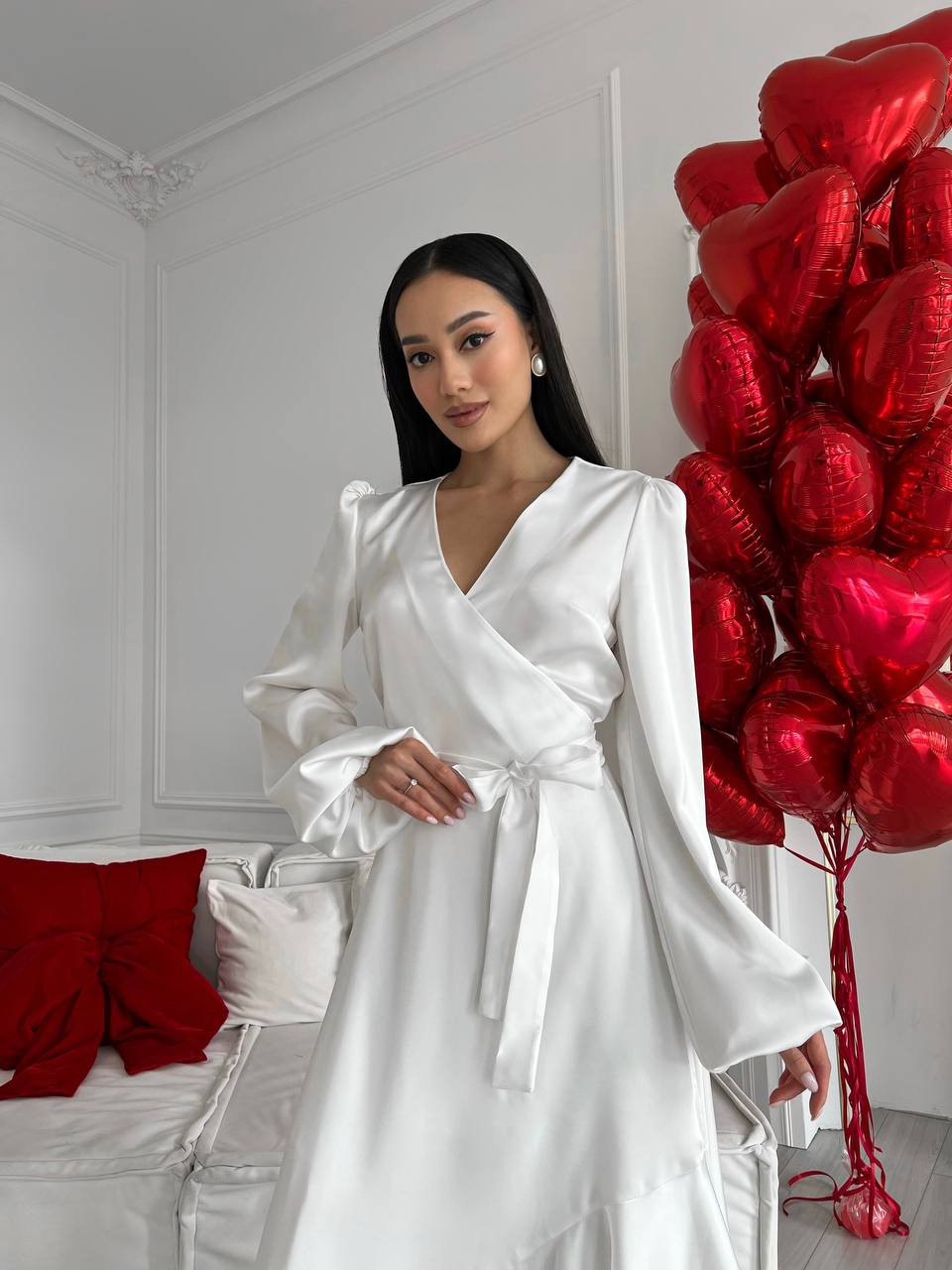 Woman in a white dress standing next to red heart-shaped balloons in a room.