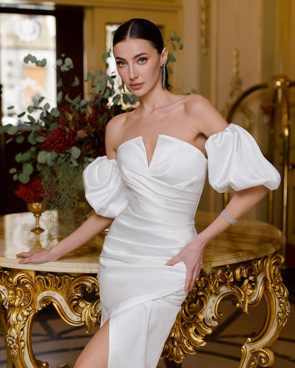 Woman in a white off-shoulder dress with puffed sleeves standing in an elegant interior setting.