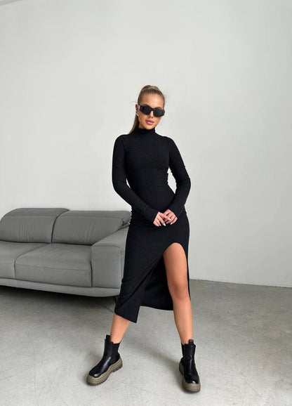 Woman wearing a black dress with a slit standing in a minimalistic room.