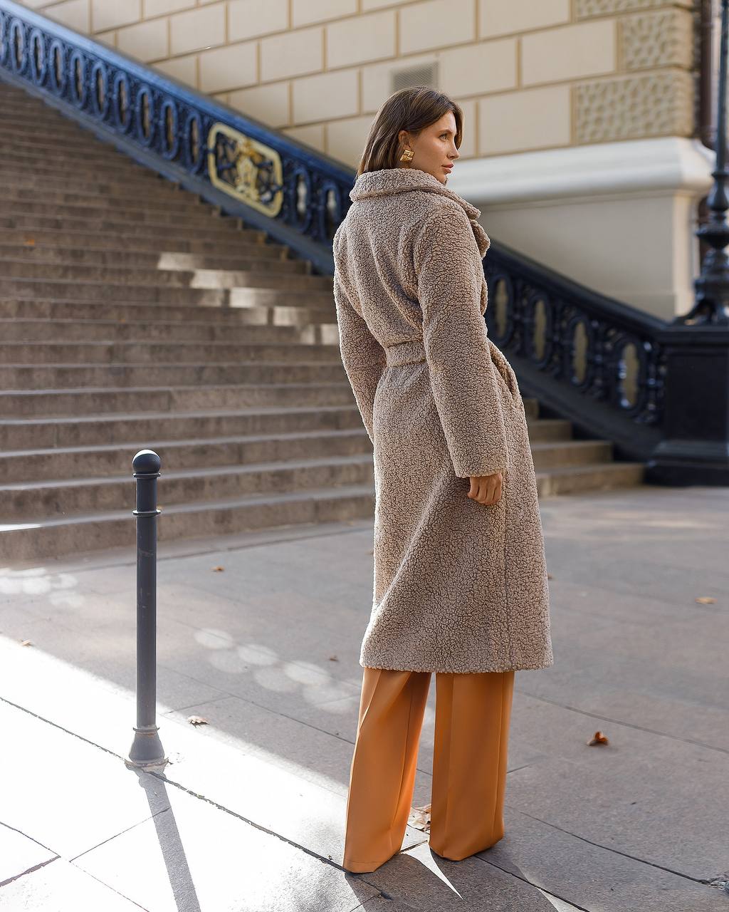 Woman wearing a long beige coat and orange pants walking on a city street.