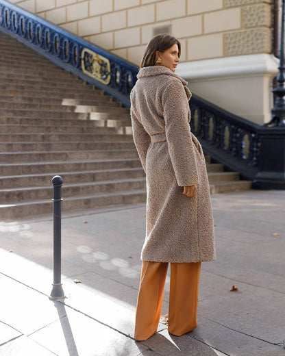 Woman wearing a long beige coat and orange pants walking on a city street.