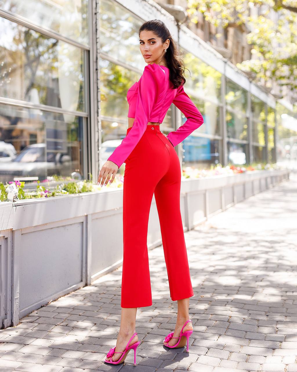Woman in a bright pink top and red pants standing on a sidewalk with a building and plants in the background.