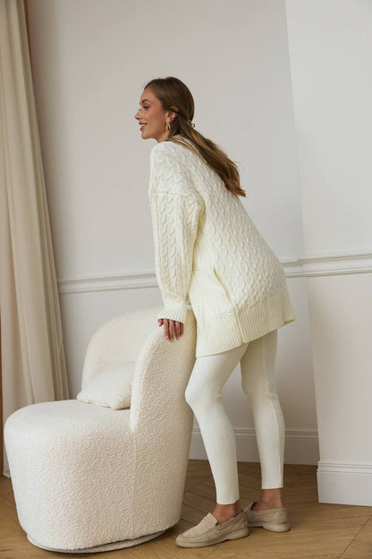 Woman in a cream-colored sweater and leggings standing next to a white chair in a room with beige curtains.