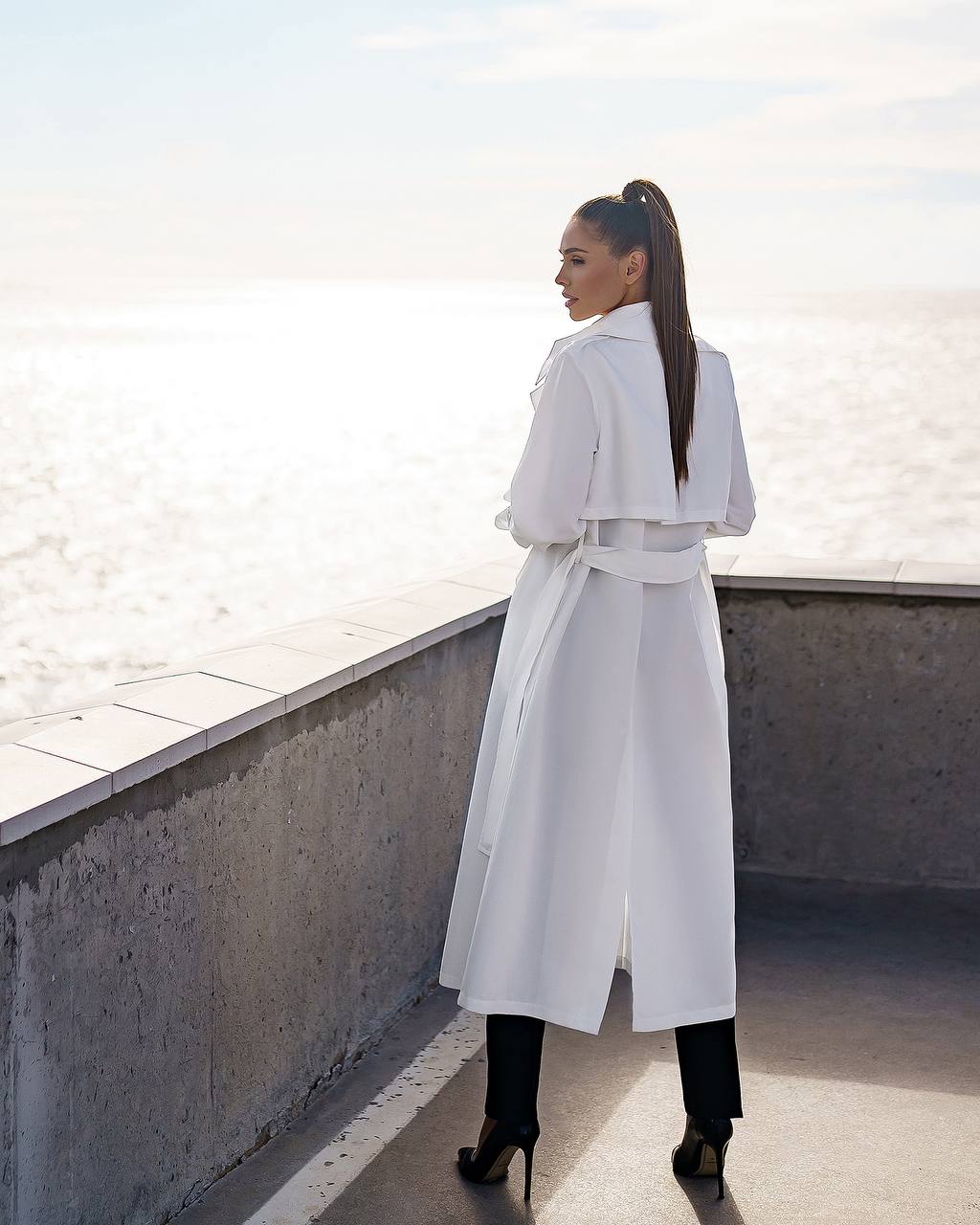 Woman in a white trench coat standing on a rooftop with a light-colored wall in the background