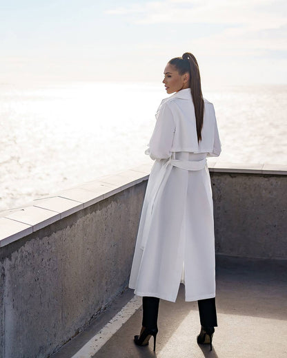 Woman in a white trench coat standing on a rooftop with a light-colored wall in the background