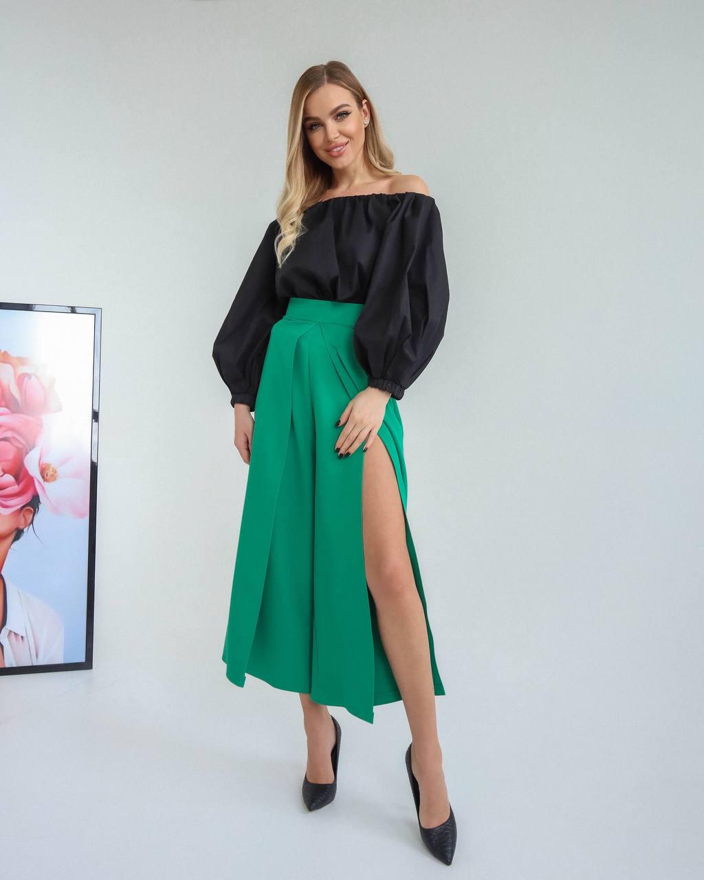 Woman wearing a black off-shoulder top and green skirt with a slit, standing against a white background.