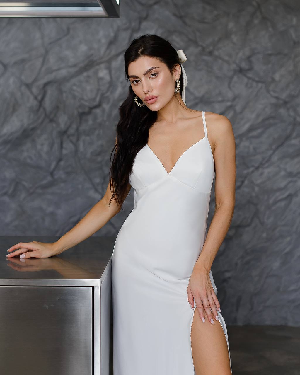 Woman in a white dress standing against a textured gray wall.