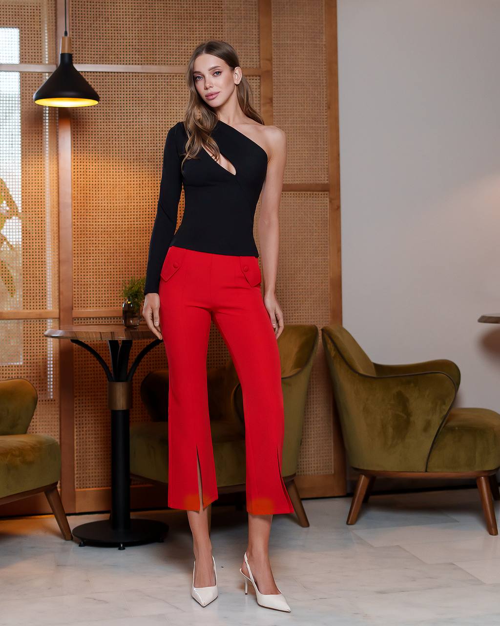 Woman in black top and red pants standing in a stylish room with green chairs and a lamp.
