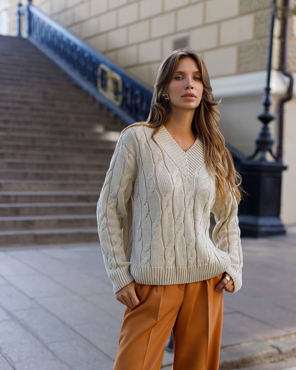 Woman wearing a beige cable knit sweater and orange pants standing on a street.