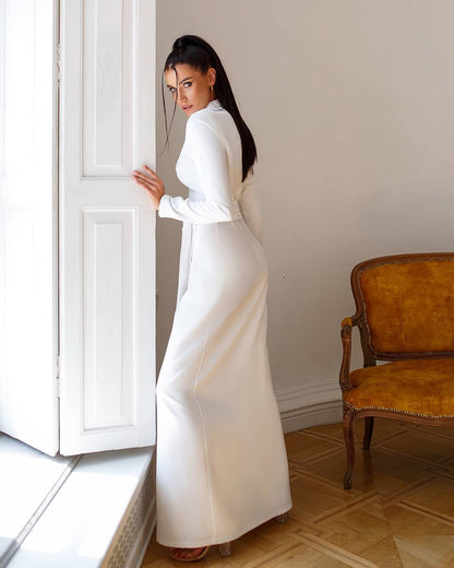 Woman in a white dress standing in a room with a chair and door.