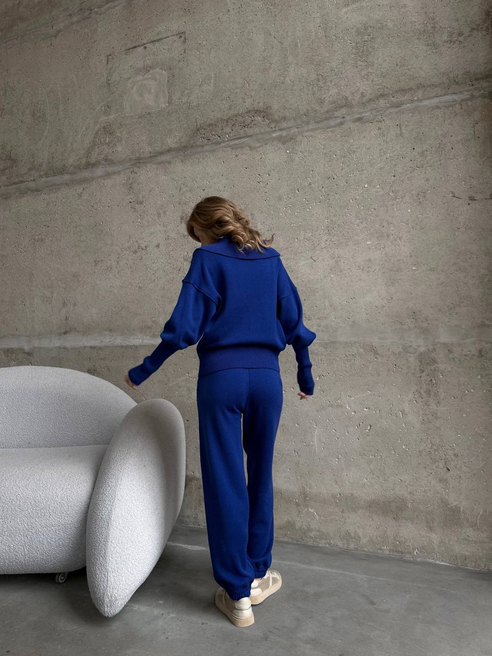 Person in a blue outfit standing in a minimalistic room with a textured wall and white chair.