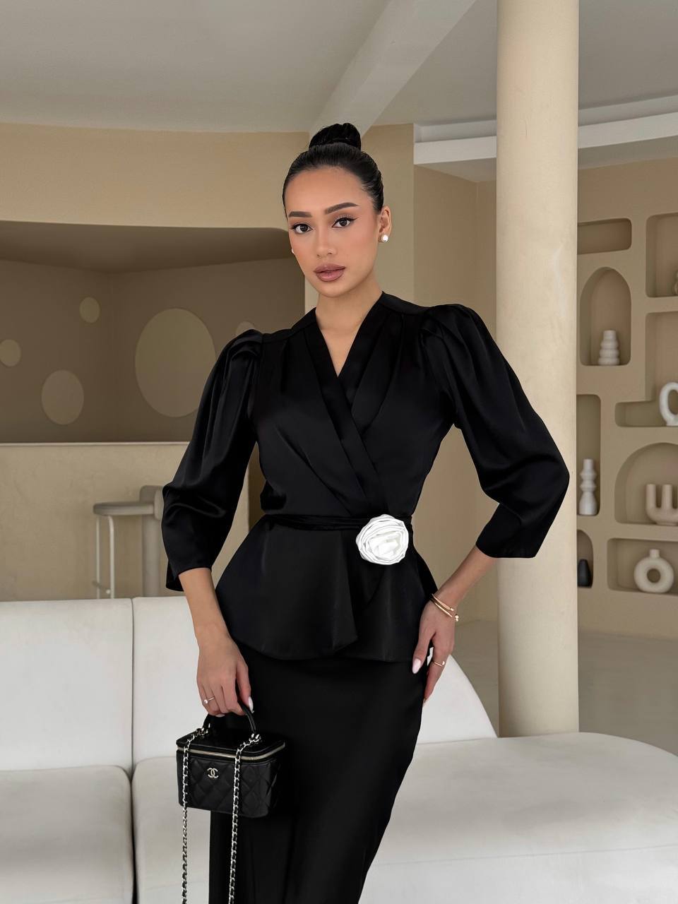 Woman in a black outfit with a white flower detail, holding a black handbag in an indoor setting.