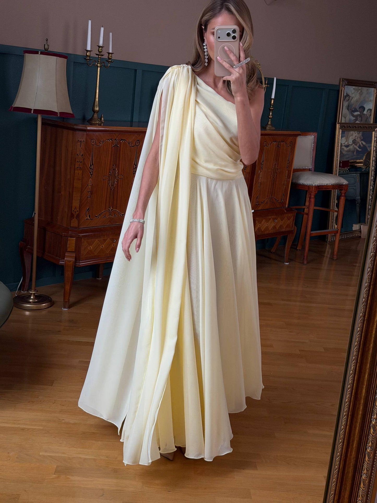 Woman in a white saree standing in a room with wooden furniture and a lamp.