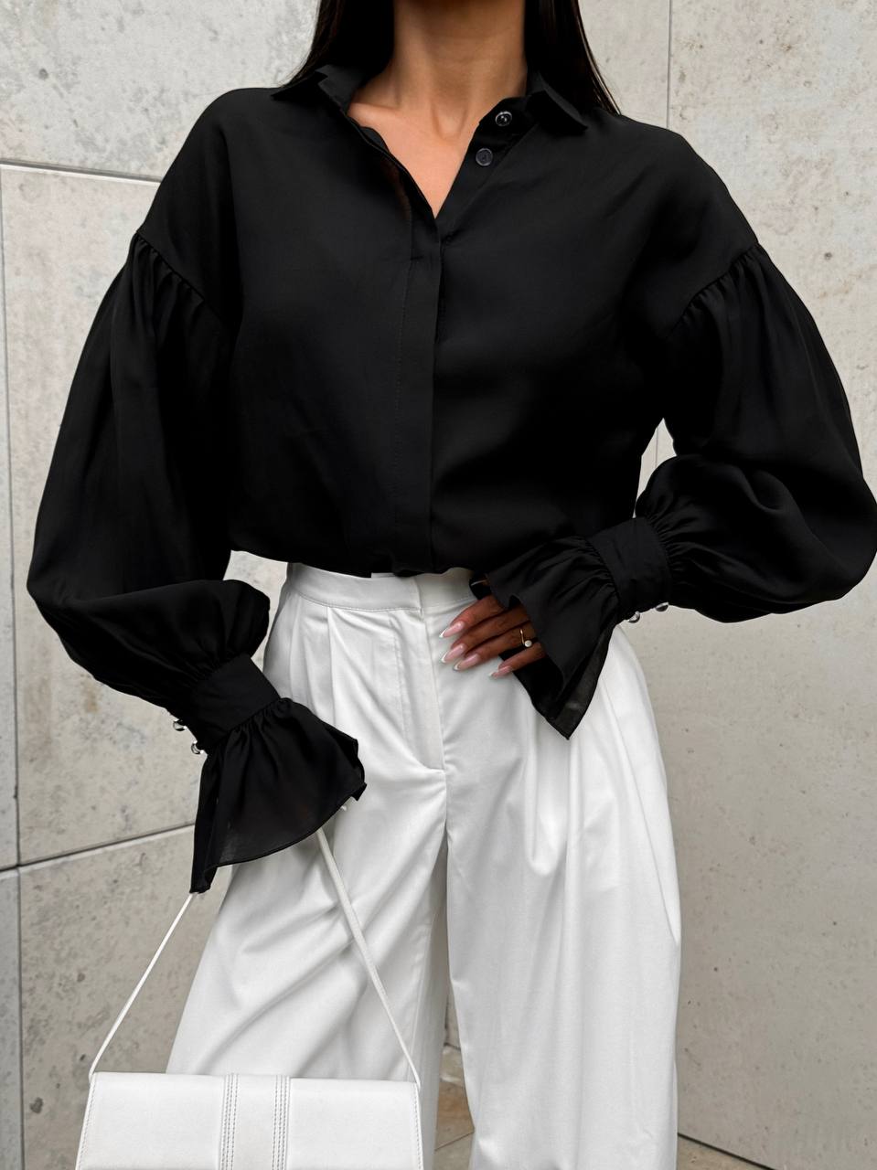 Person wearing a black blouse with puffed sleeves and white pants against a gray wall.