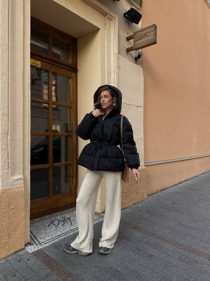 Person wearing a black puffer jacket and white pants standing on a sidewalk.