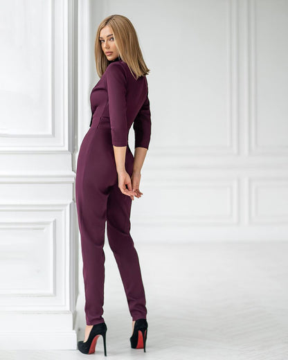 Woman wearing a purple jumpsuit standing against a white paneled wall.