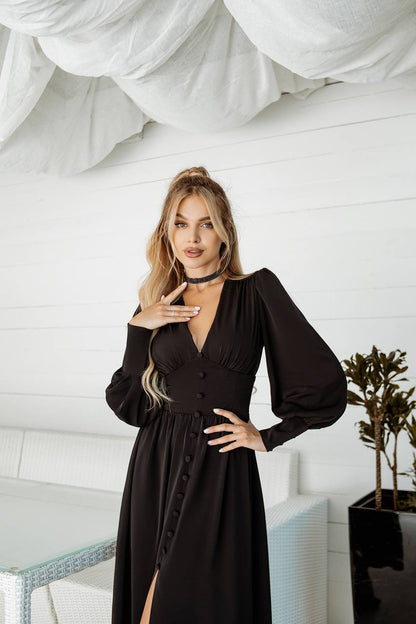 Woman in a black dress posing in a room with white walls and a plant.