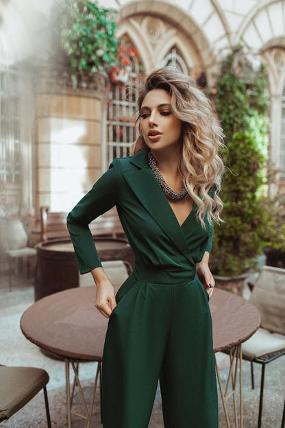 Woman in a green jumpsuit standing in an indoor setting with plants and tables.
