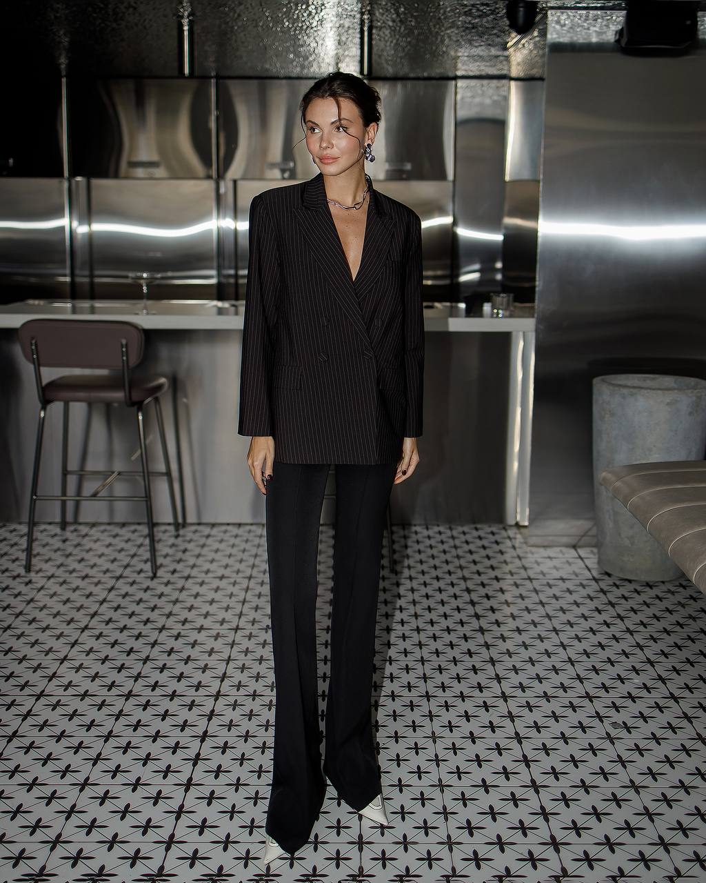 Woman in a black pinstripe suit standing in a modern interior setting with metallic walls and patterned floor.