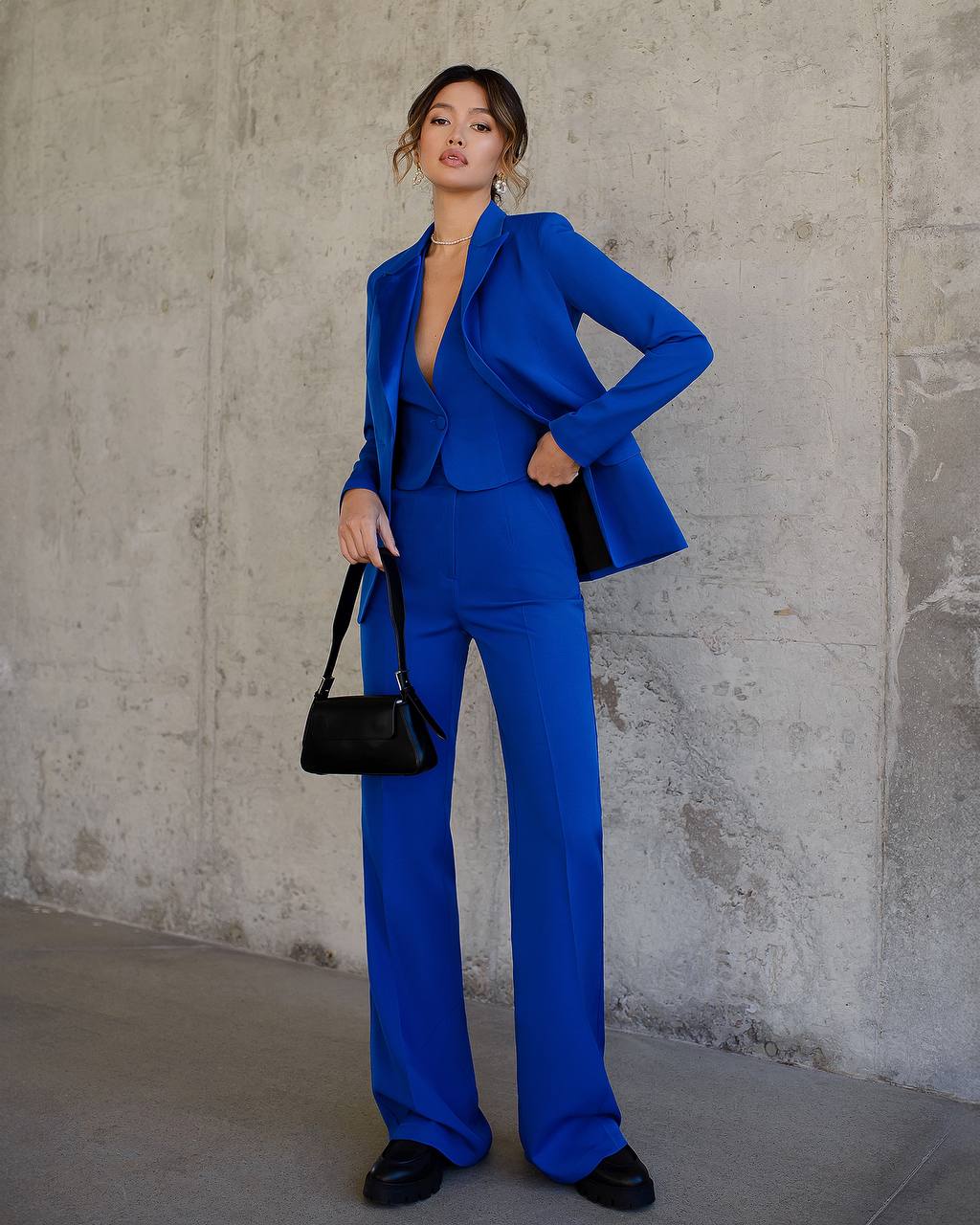 Woman in a blue suit holding a black handbag against a gray concrete wall.