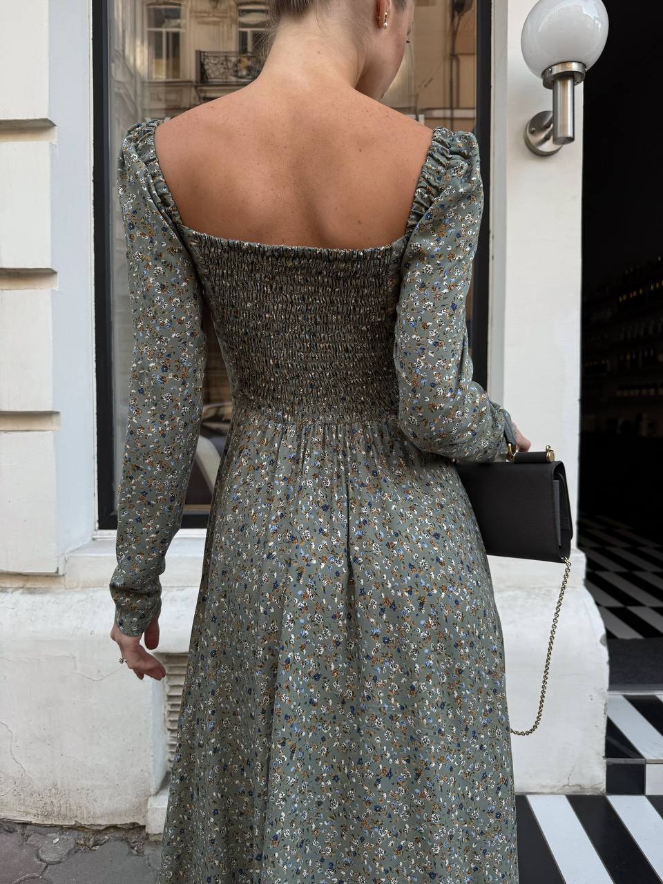 Woman wearing a floral dress with long sleeves, standing in front of a building entrance.