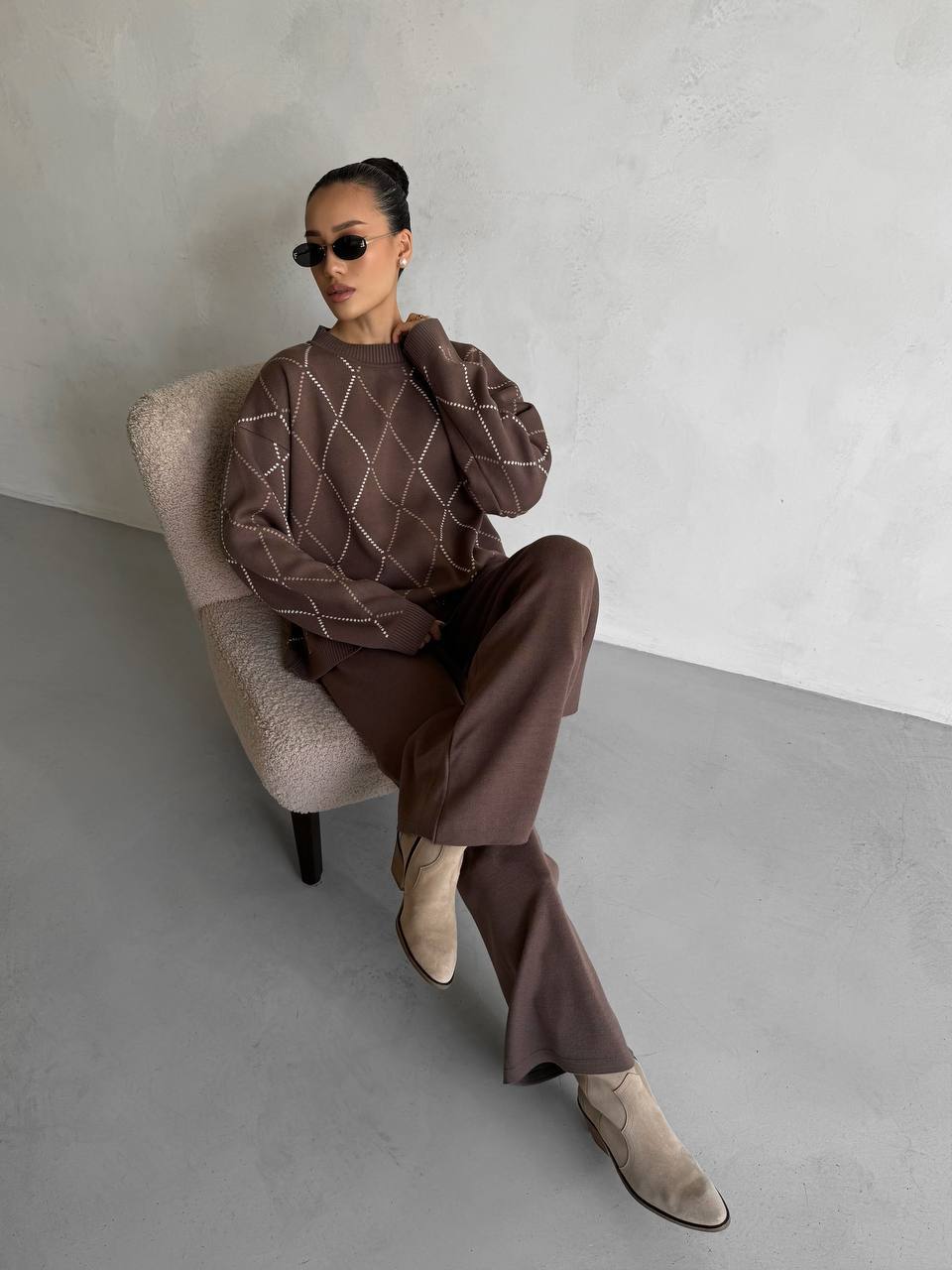 Woman sitting on a chair wearing a brown outfit with sunglasses against a plain background