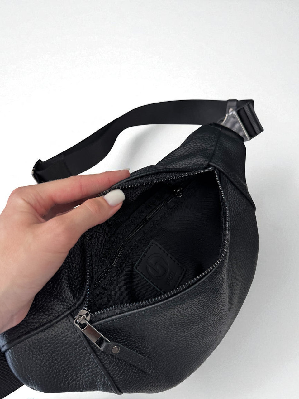 A black pebbled leather crossbody bag shown open, with a hand holding the zipper flap to reveal the black interior lining and a small embossed logo patch inside.